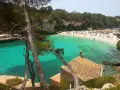 Piękne majorka plaże z turkusową wodą, skalistymi klifami i sosnami. W oddali widać tłum plażowiczów i mały domek z ceglanym dachem.