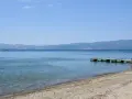 Spokojne, błękitne wody jeziora, piaszczyste plaże z kamieniami i drewniane molo. W oddali góry.