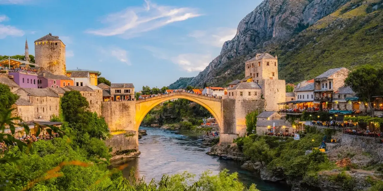 Mostar most, kamienny łuk nad rzeką Neretwą, otoczony starymi budynkami i zielenią, oświetlony zachodzącym słońcem.