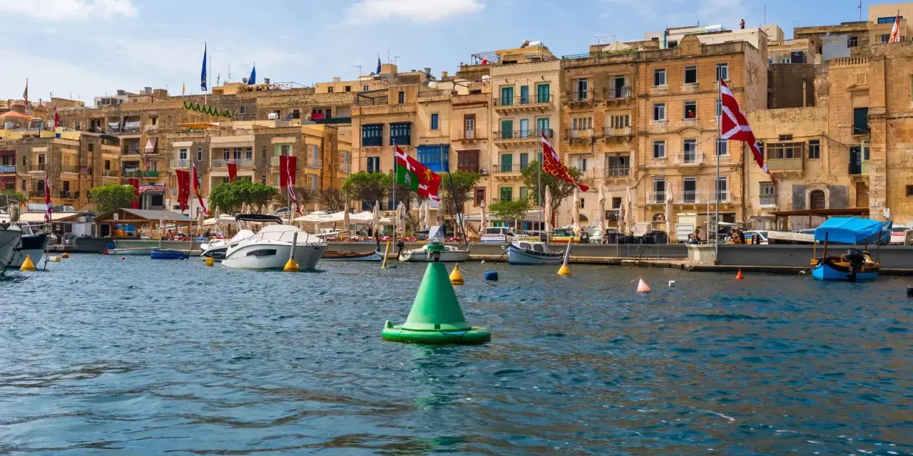 Malta w sierpniu: port z łodziami i kamiennymi budynkami, ozdobiony flagami.