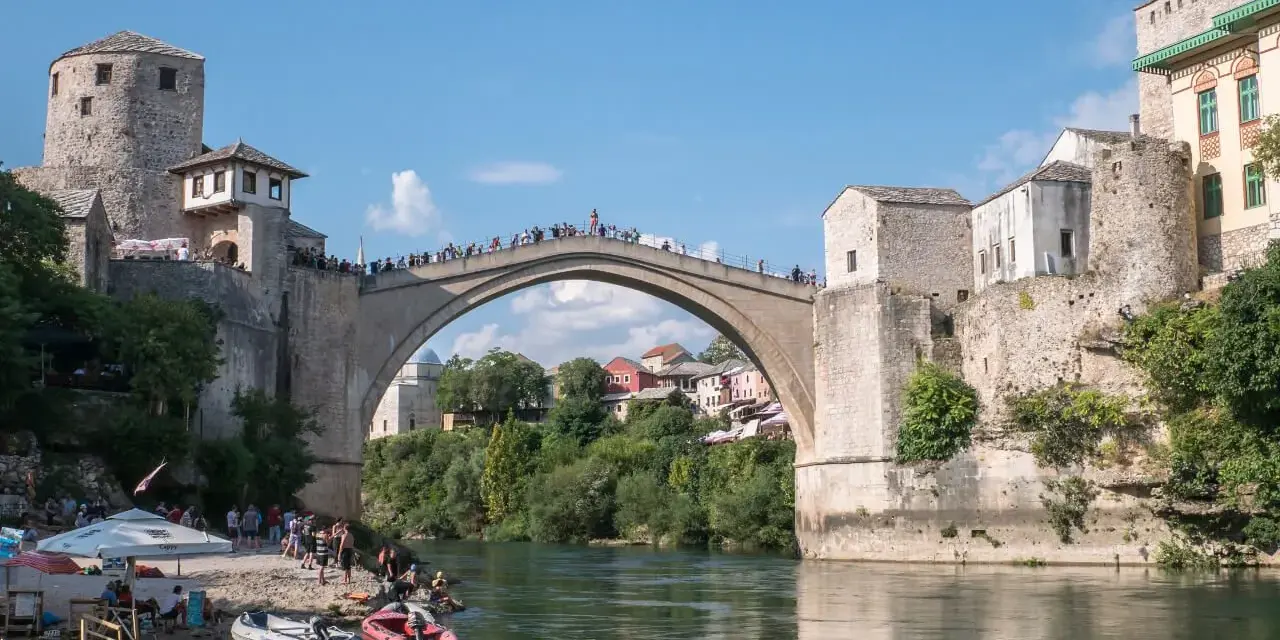 Mostar, Bośnia i Hercegowina: Słynny Stary Most z kamienia, tłumy ludzi na moście i brzegu, łodzie na rzece Neretwie.