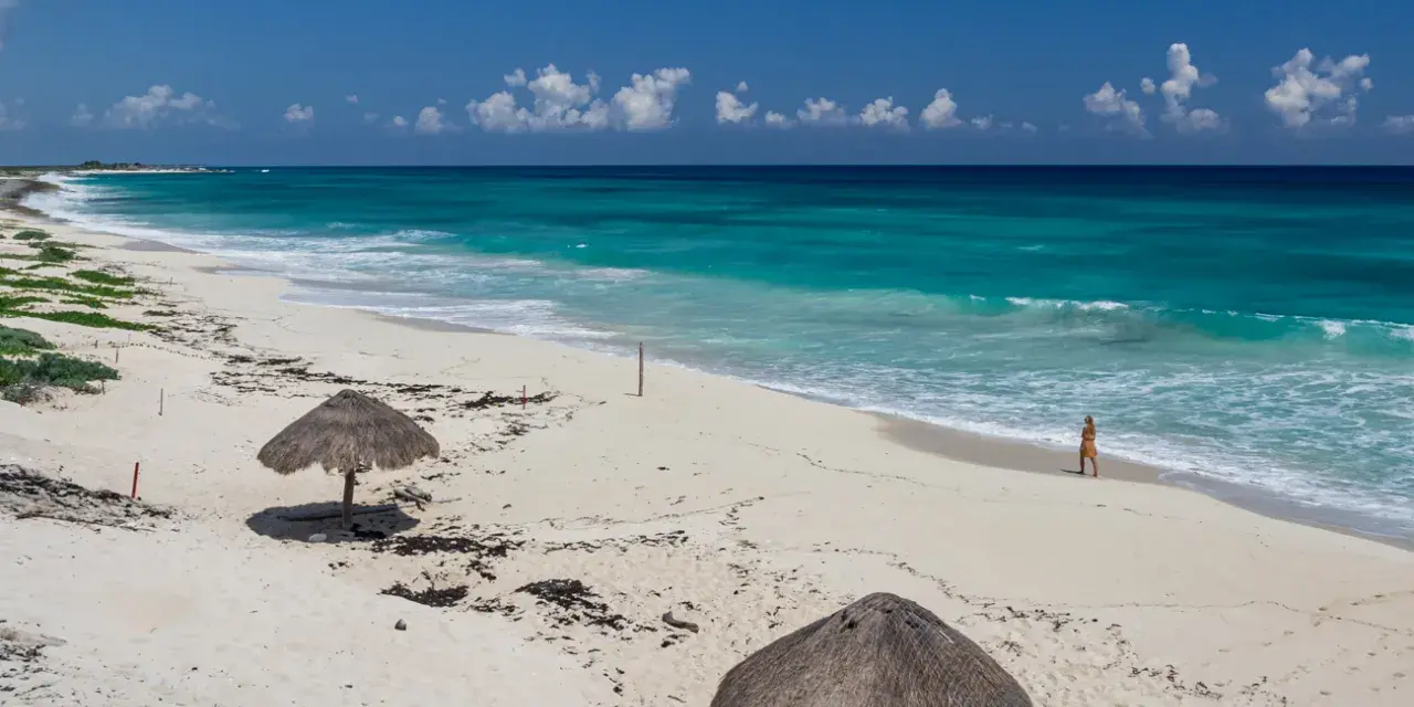Pusta, piaszczysta nordeste playa z turkusową wodą i dwoma słomianymi parasolami. Kobieta spaceruje brzegiem morza.