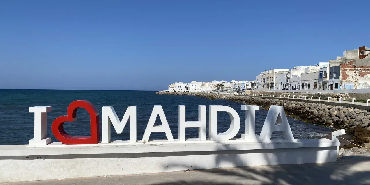 Napis "I ❤️ MAHDIA" na tle morza i białych budynków. Słońce i cień palm. Tunezja Mahdia.