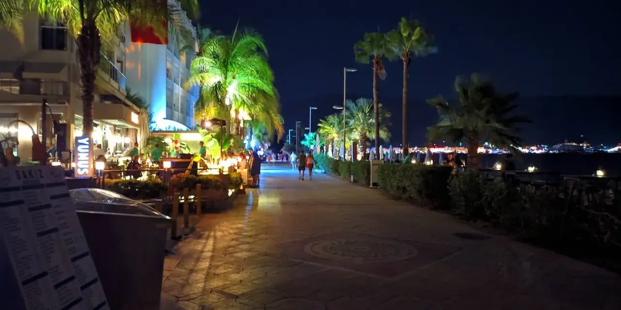 Wieczorny spacer promenadą w Bułgarii we wrześniu. Palmy oświetlone, restauracje tętnią życiem, a w oddali widać światła miasta.