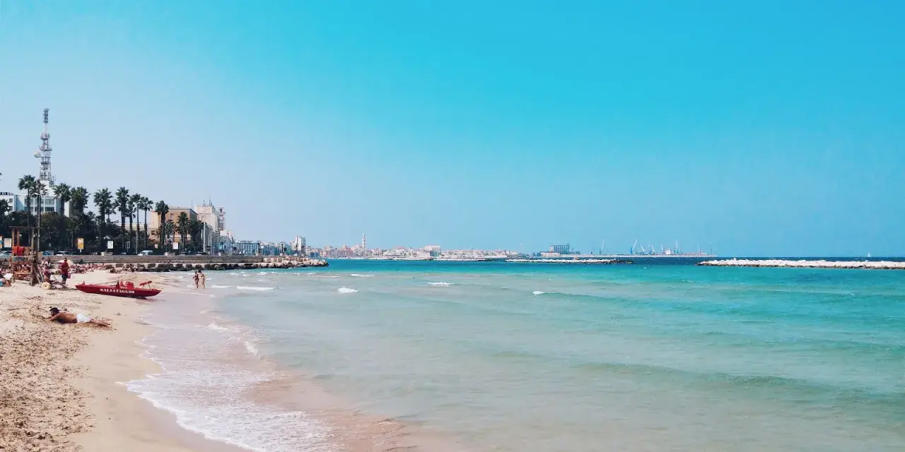 Słoneczne bari plaże z turkusową wodą, palmami i miastem w tle. Czerwona łódź ratownicza czeka na plaży.