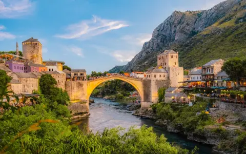 Mostar most, kamienny łuk nad rzeką Neretwą, otoczony starymi budynkami i zielenią, oświetlony zachodzącym słońcem.