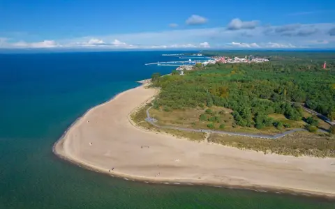 Plaża i las na Helu. Widok z lotu ptaka na turkusowe morze, piaszczystą plażę i zielony las. W oddali widać zabudowania i port. Hel atrakcje czekają!