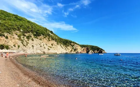 Słoneczne plaże w Budvie, z turkusową wodą i zielonymi wzgórzami w tle. Ludzie relaksują się na kamienistym brzegu i pływają w morzu.