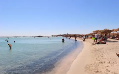 Słoneczna plaża Elafonisi z turkusową wodą i piaskiem, pełna ludzi kąpiących się i odpoczywających pod parasolami.