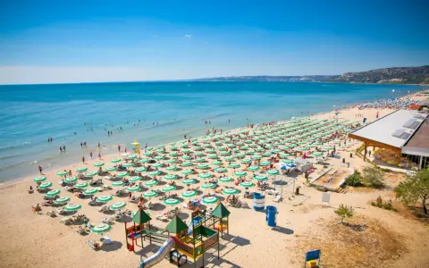 Bułgaria, Złote Piaski: rozległa plaża z rzędami zielonych parasoli, turystami kąpiącymi się w morzu i placem zabaw. Atrakcje dla każdego!