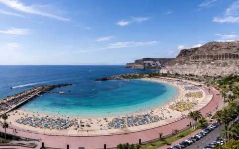 Słoneczna plaża z turkusową wodą i mnóstwem leżaków. W oddali klify i hotele, idealne gran canaria atrakcje dla każdego.