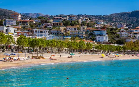 Albania, pogoda czerwiec: słoneczny dzień na plaży, turkusowe morze, piaszczysty brzeg, zielone drzewa i białe budynki na wzgórzu.