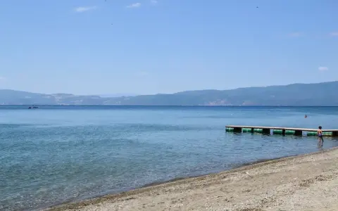 Spokojne, błękitne wody jeziora, piaszczyste plaże z kamieniami i drewniane molo. W oddali góry.