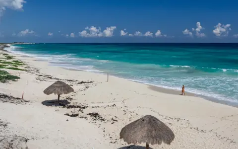 Pusta, piaszczysta nordeste playa z turkusową wodą i dwoma słomianymi parasolami. Kobieta spaceruje brzegiem morza.