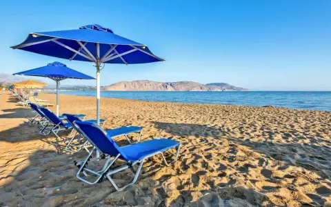 Błękitne leżaki i parasole na piaszczystej plaży Kavros Beach, z widokiem na spokojne morze i górzyste wybrzeże w oddali.