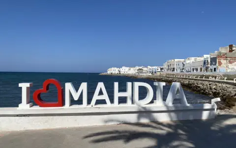 Napis "I ❤️ MAHDIA" na tle morza i białych budynków. Słońce i cień palm. Tunezja Mahdia.