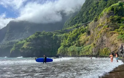 Surfer z niebieską deską na seixal beach, w tle zielone klify i chmury.