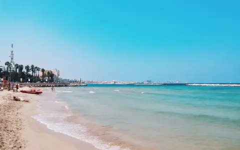 Słoneczne bari plaże z turkusową wodą, palmami i miastem w tle. Czerwona łódź ratownicza czeka na plaży.