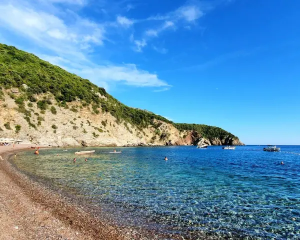 Słoneczne plaże w Budvie, z turkusową wodą i zielonymi wzgórzami w tle. Ludzie relaksują się na kamienistym brzegu i pływają w morzu.