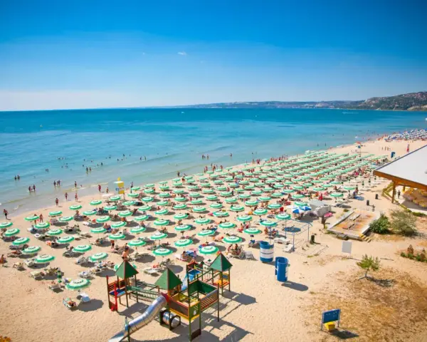 Bułgaria, Złote Piaski: rozległa plaża z rzędami zielonych parasoli, turystami kąpiącymi się w morzu i placem zabaw. Atrakcje dla każdego!