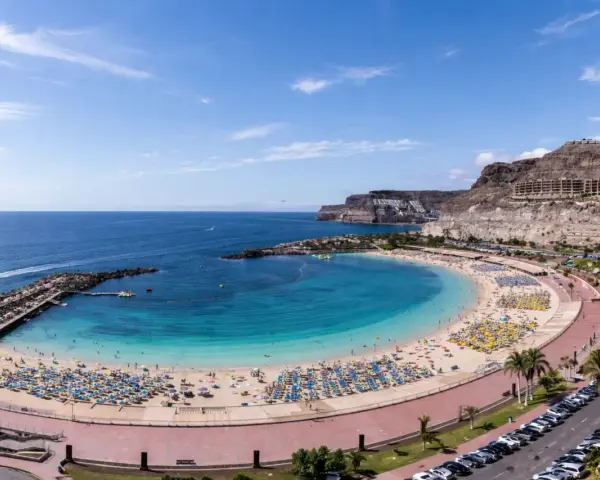 Słoneczna plaża z turkusową wodą i mnóstwem leżaków. W oddali klify i hotele, idealne gran canaria atrakcje dla każdego.