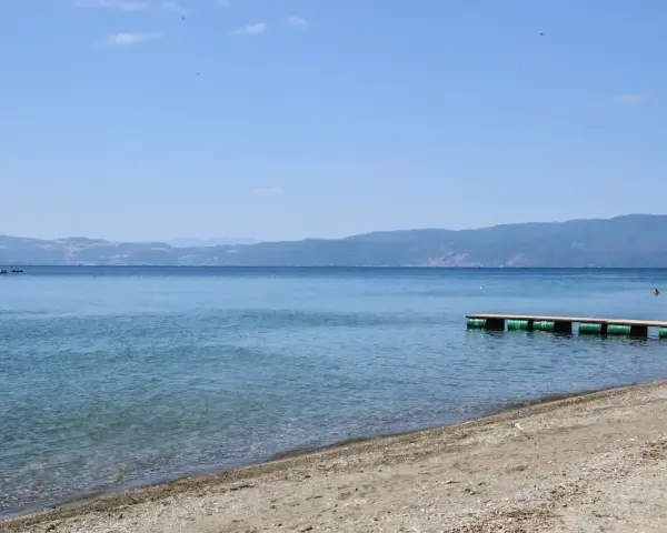 Spokojne, błękitne wody jeziora, piaszczyste plaże z kamieniami i drewniane molo. W oddali góry.