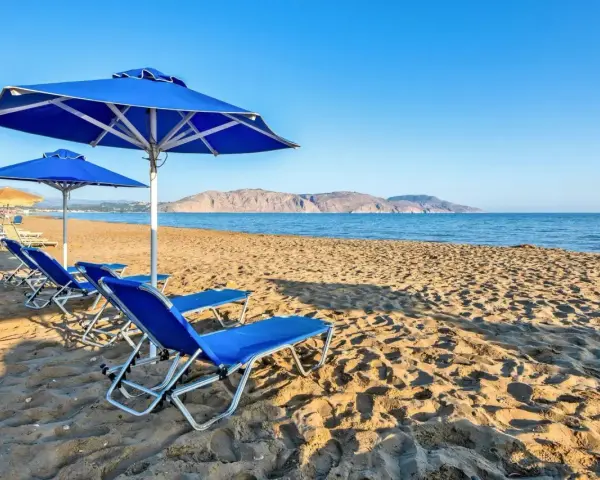 Błękitne leżaki i parasole na piaszczystej plaży Kavros Beach, z widokiem na spokojne morze i górzyste wybrzeże w oddali.