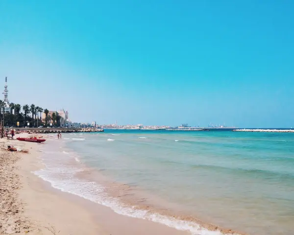 Słoneczne bari plaże z turkusową wodą, palmami i miastem w tle. Czerwona łódź ratownicza czeka na plaży.