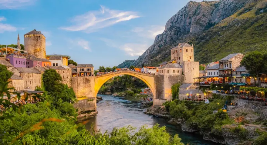Mostar most, kamienny łuk nad rzeką Neretwą, otoczony starymi budynkami i zielenią, oświetlony zachodzącym słońcem.