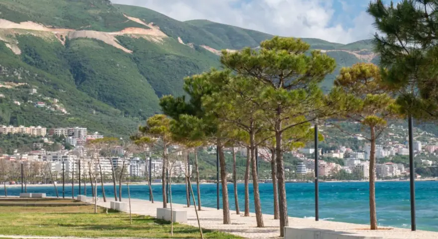 Wlora, Albania: nadmorska promenada z drzewami i widokiem na miasto u podnóża zielonych wzgórz.