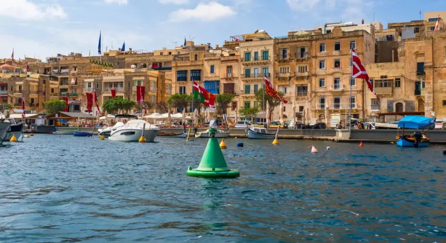 Malta w sierpniu: port z łodziami i kamiennymi budynkami, ozdobiony flagami.
