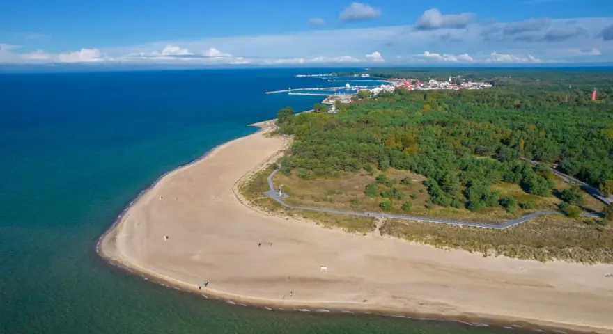Plaża i las na Helu. Widok z lotu ptaka na turkusowe morze, piaszczystą plażę i zielony las. W oddali widać zabudowania i port. Hel atrakcje czekają!