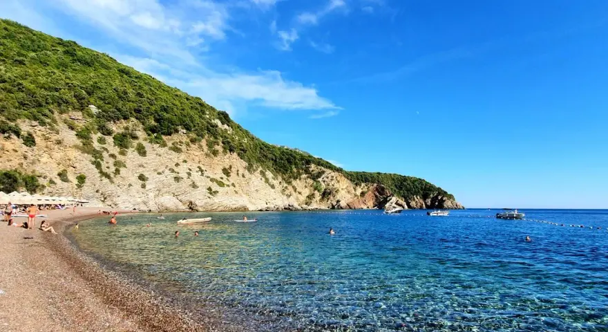Słoneczne plaże w Budvie, z turkusową wodą i zielonymi wzgórzami w tle. Ludzie relaksują się na kamienistym brzegu i pływają w morzu.
