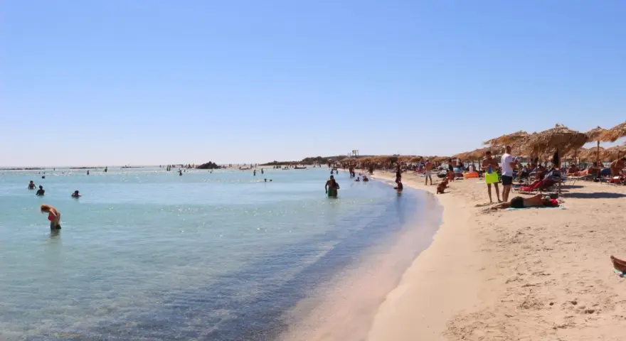 Słoneczna plaża Elafonisi z turkusową wodą i piaskiem, pełna ludzi kąpiących się i odpoczywających pod parasolami.