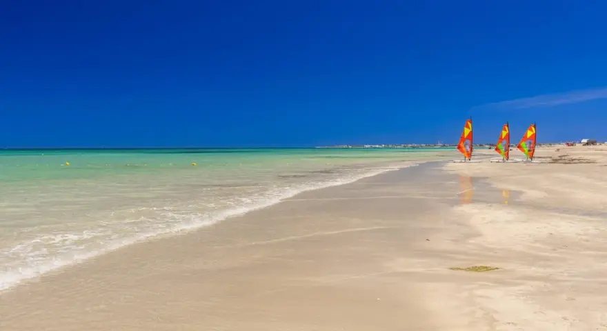 Piękna, słoneczna plaża w Tunezji we wrześniu. Trzy kolorowe żagle desek do windsurfingu stoją na piasku.