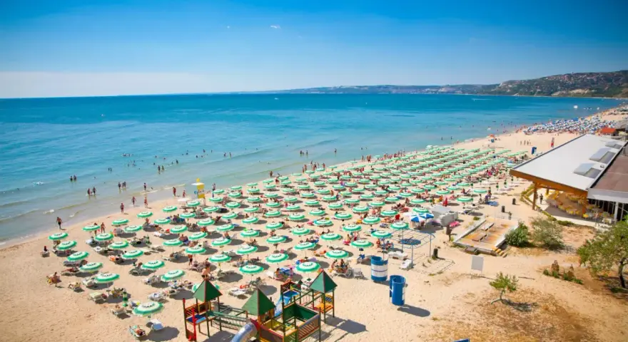 Bułgaria, Złote Piaski: rozległa plaża z rzędami zielonych parasoli, turystami kąpiącymi się w morzu i placem zabaw. Atrakcje dla każdego!