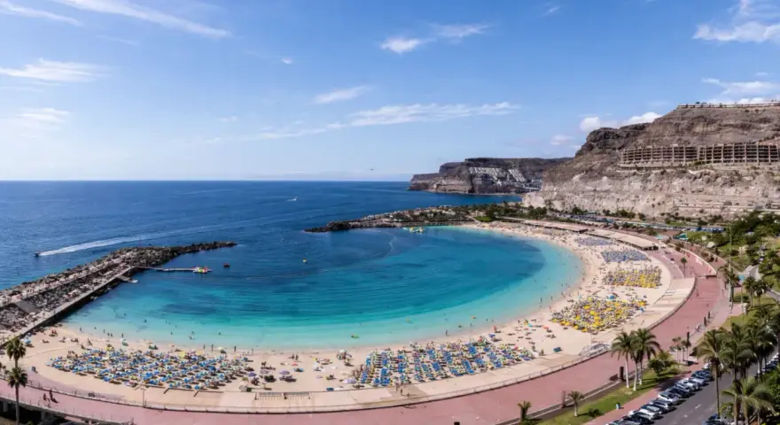 Słoneczna plaża z turkusową wodą i mnóstwem leżaków. W oddali klify i hotele, idealne gran canaria atrakcje dla każdego.