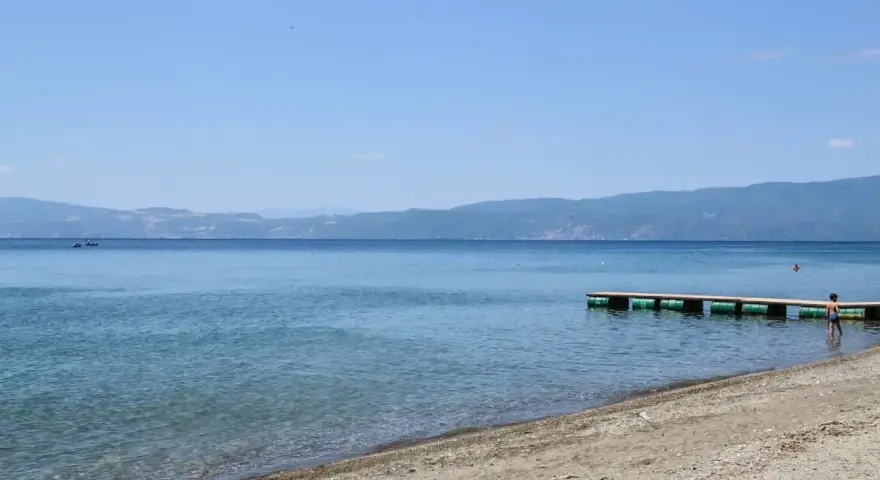 Spokojne, błękitne wody jeziora, piaszczyste plaże z kamieniami i drewniane molo. W oddali góry.