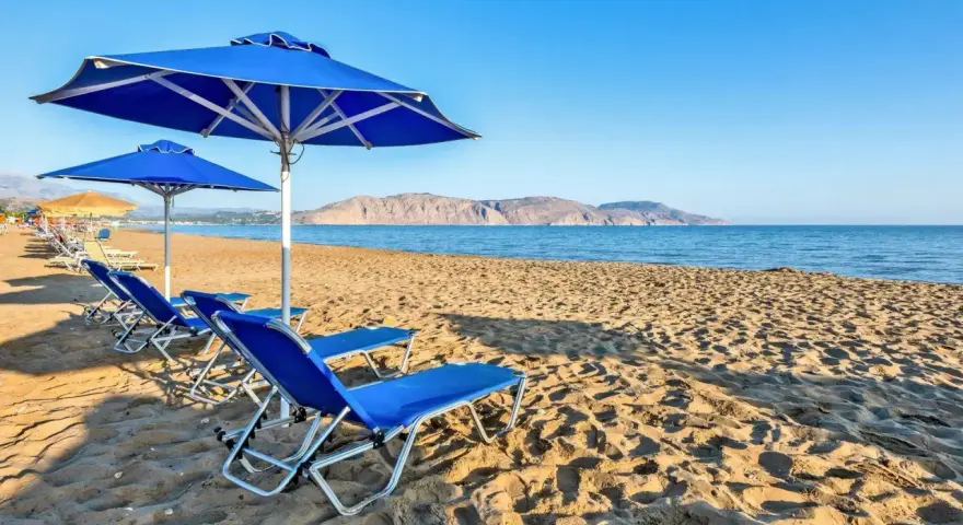Błękitne leżaki i parasole na piaszczystej plaży Kavros Beach, z widokiem na spokojne morze i górzyste wybrzeże w oddali.