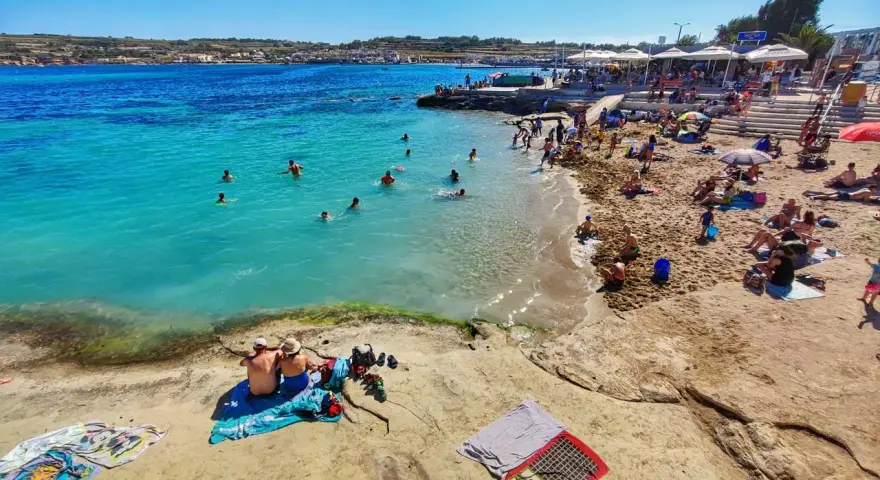 Słoneczna plaża na Malcie, ludzie kąpią się w turkusowej wodzie. Idealna temperatura do opalania i pływania.