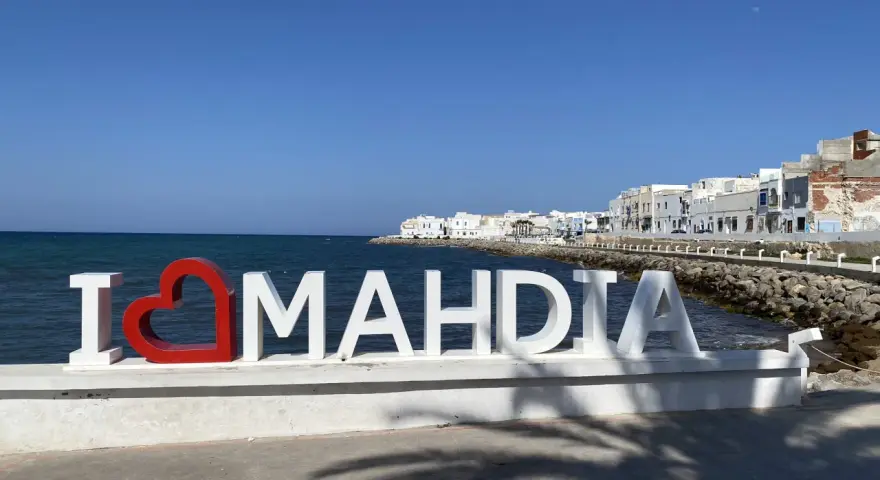 Napis "I ❤️ MAHDIA" na tle morza i białych budynków. Słońce i cień palm. Tunezja Mahdia.