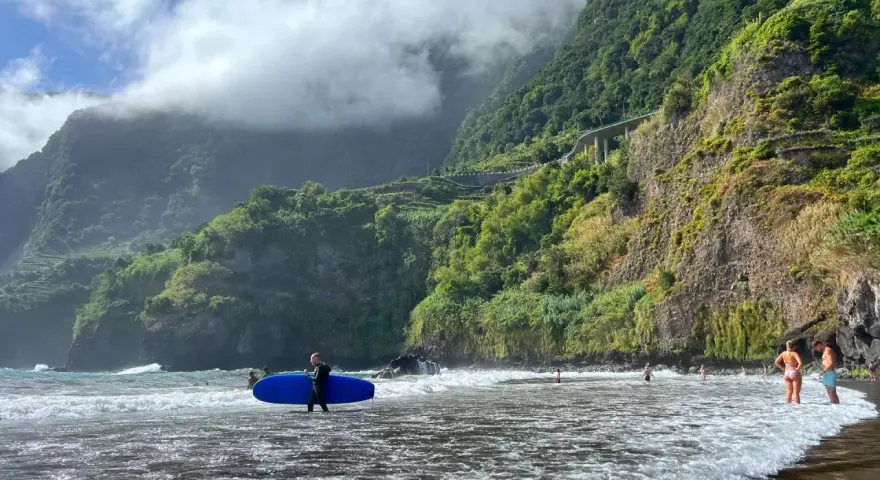 Surfer z niebieską deską na seixal beach, w tle zielone klify i chmury.