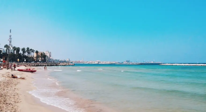 Słoneczne bari plaże z turkusową wodą, palmami i miastem w tle. Czerwona łódź ratownicza czeka na plaży.