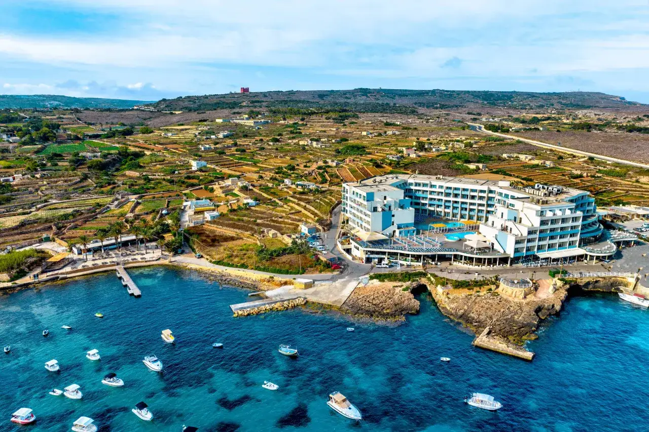 Malta w sierpniu: hotel z basenem i tarasami nad turkusowym morzem, w tle tarasowe pola i wieża.