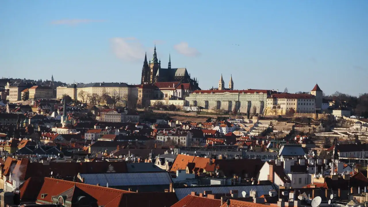 Widok na praskie dachy i majestatyczny zamek na Hradczanach, górujący nad miastem w słoneczny dzień.