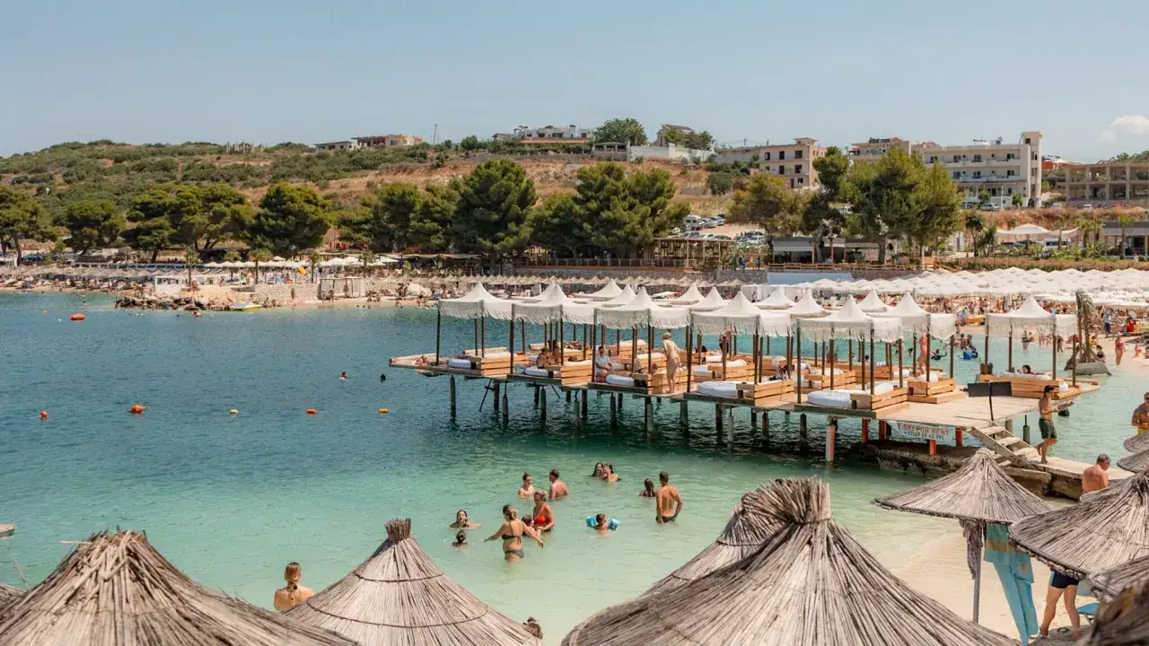 Wakacje w Albanii: ludzie kąpią się w turkusowej wodzie, odpoczywają na leżakach na pomoście i plaży.
