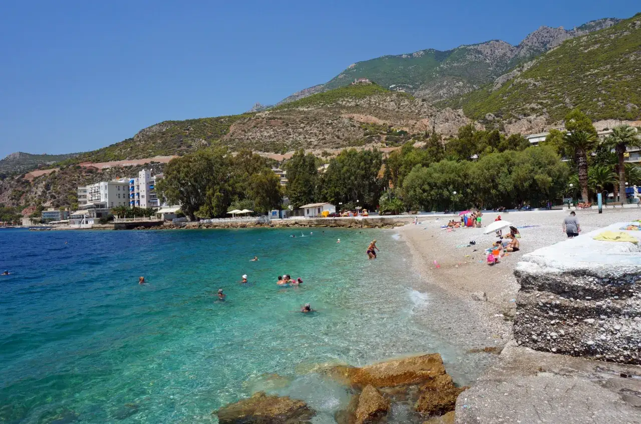 Słoneczny dzień na plaży w Loutraki, Grecja. Ludzie kąpią się w krystalicznie czystej wodzie, podczas gdy góry górują nad wybrzeżem.