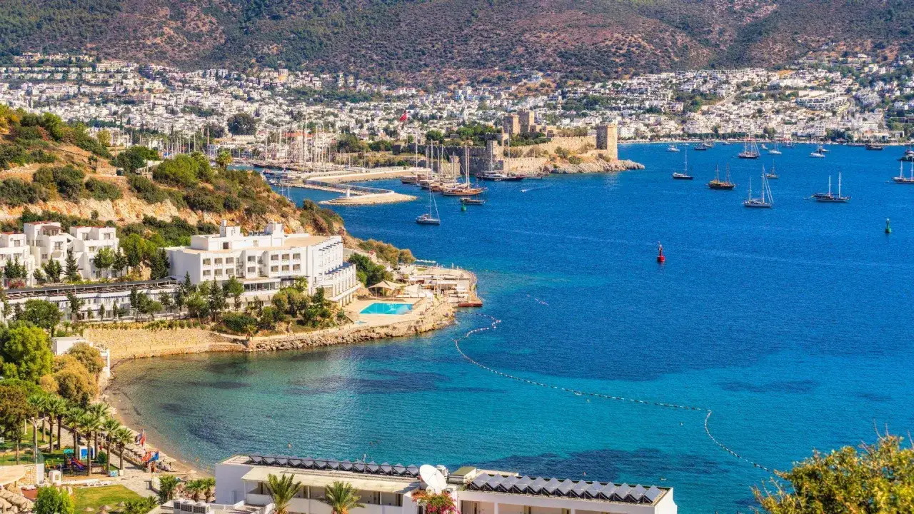 Piękna pogoda w Bodrum w Turcji. Białe budynki hotelowe, basen, plaża i zatoka pełna łodzi żaglowych. W tle zamek i góry.