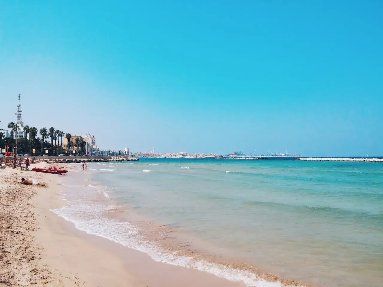Słoneczne bari plaże z turkusową wodą, palmami i miastem w tle. Czerwona ł&oacute;dź ratownicza czeka na plaży.