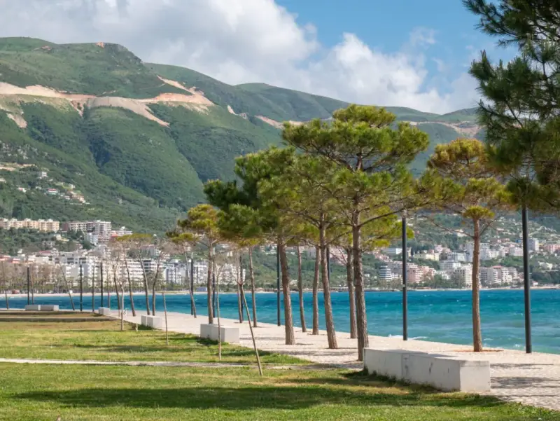 Wlora, Albania: nadmorska promenada z drzewami i widokiem na miasto u podnóża zielonych wzgórz.